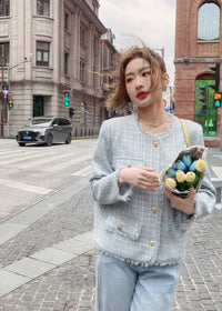 A young woman poses on a cobblestone street corner with European-style architecture in the background. She is wearing a buttoned light blue-gray tweed jacket and holding a small bouquet of blue and yellow tulips.