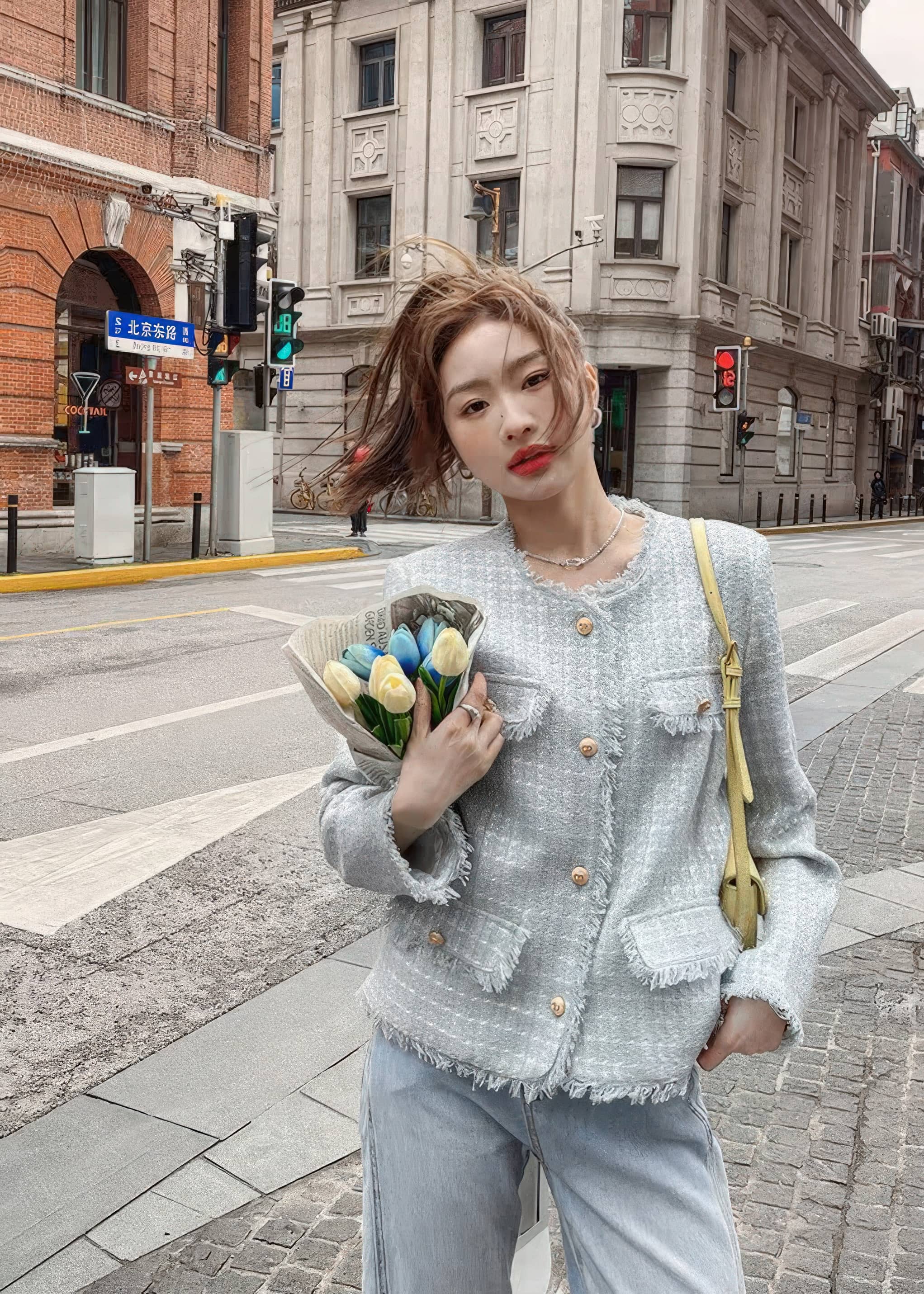 A young woman stands on an urban street holding a bouquet of tulips. She is wearing a light blue-gray tweed jacket with gold button accents and raw trim, paired with loose light-blue jeans and a yellow purse.