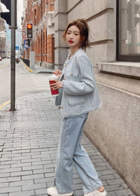 A side profile shot of a young woman walking down a city street next to a stone building. She is wearing a light blue-gray tweed jacket, wide-leg denim jeans, white shoes, and holding a takeaway coffee cup.