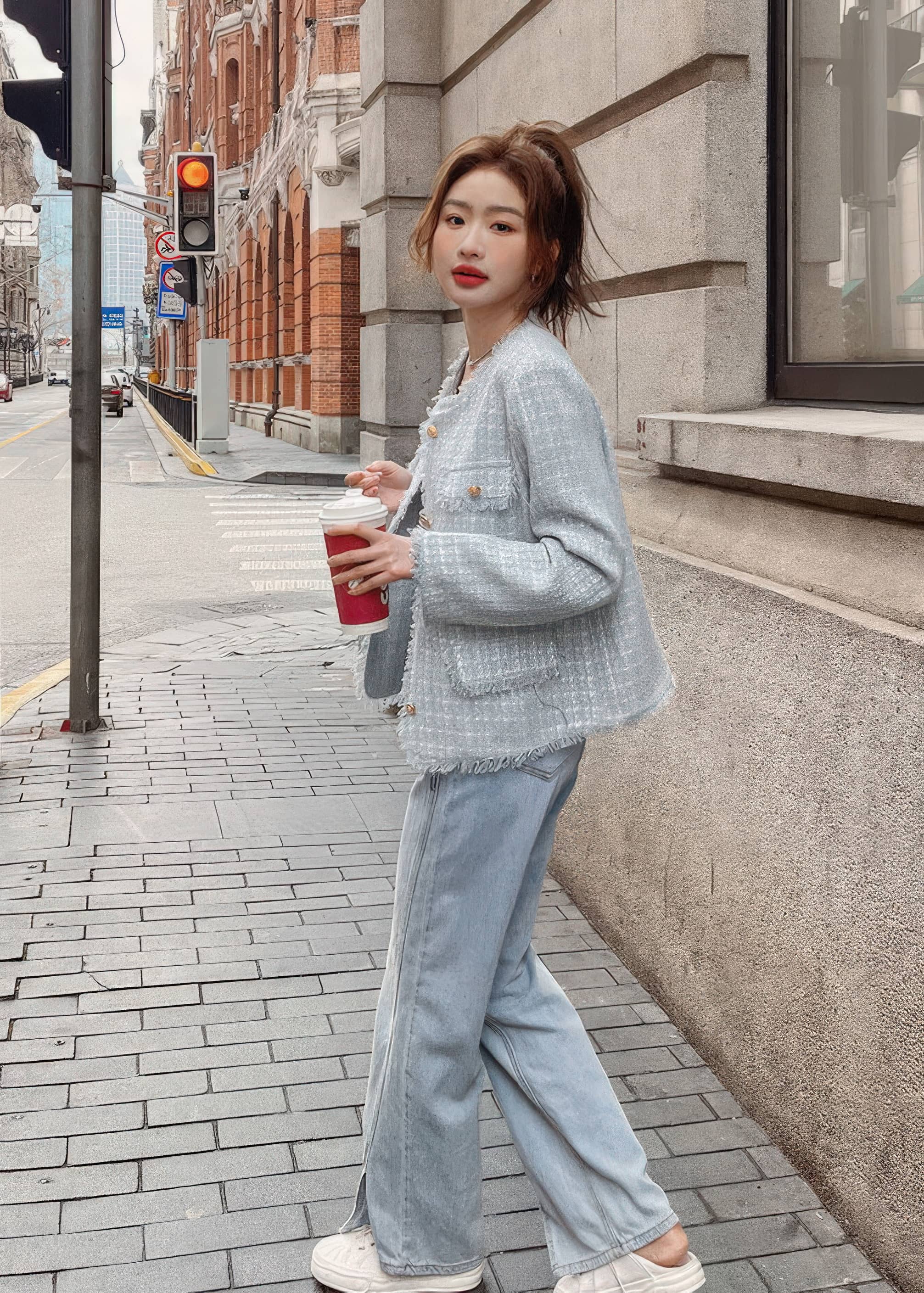 A side profile shot of a young woman walking down a city street next to a stone building. She is wearing a light blue-gray tweed jacket, wide-leg denim jeans, white shoes, and holding a takeaway coffee cup.