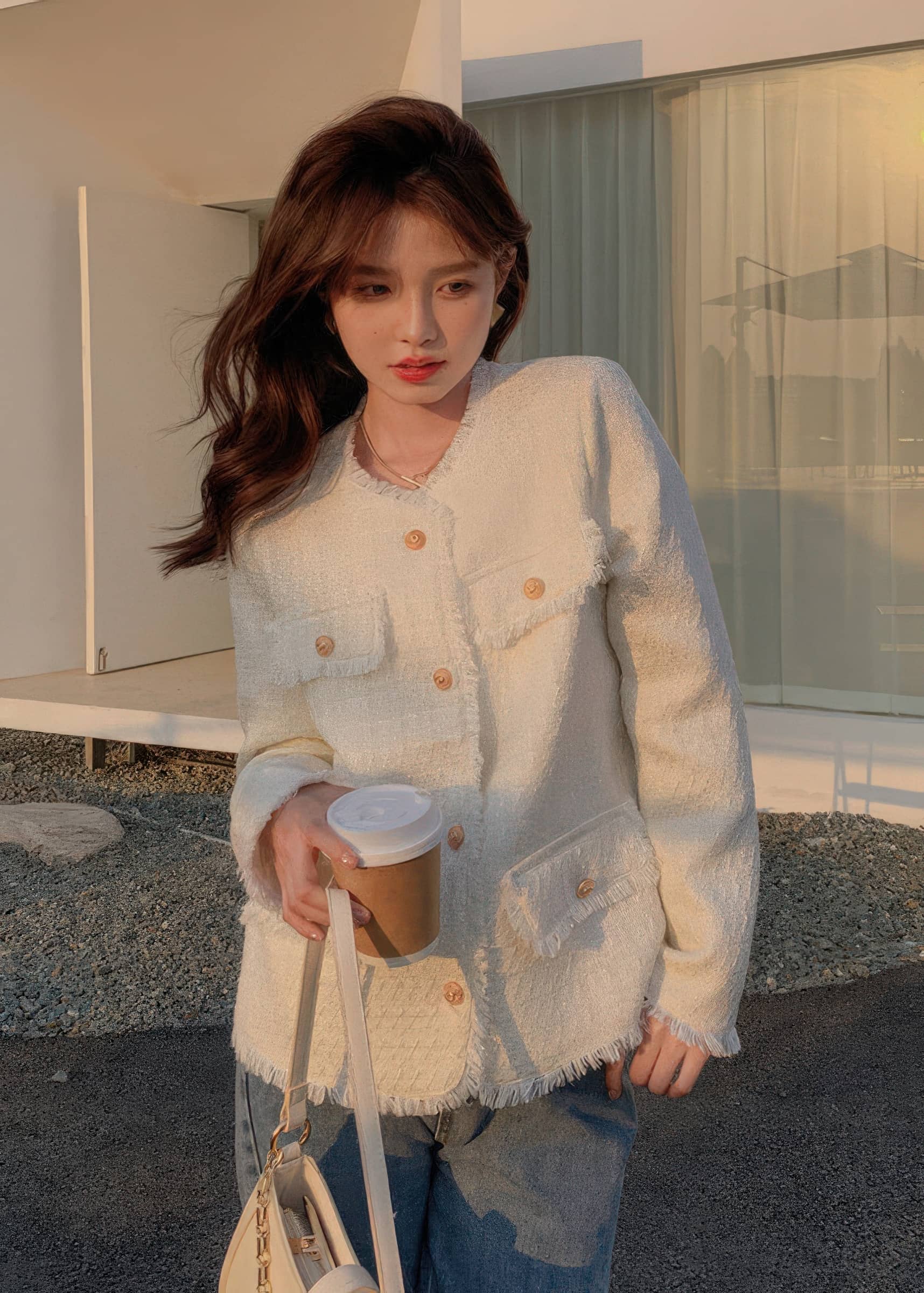 A young woman with long, wavy brown hair poses outdoors during golden hour, wearing a structured white tweed jacket buttoned halfway over a white top. She is holding a coffee cup and a cream-colored shoulder bag.