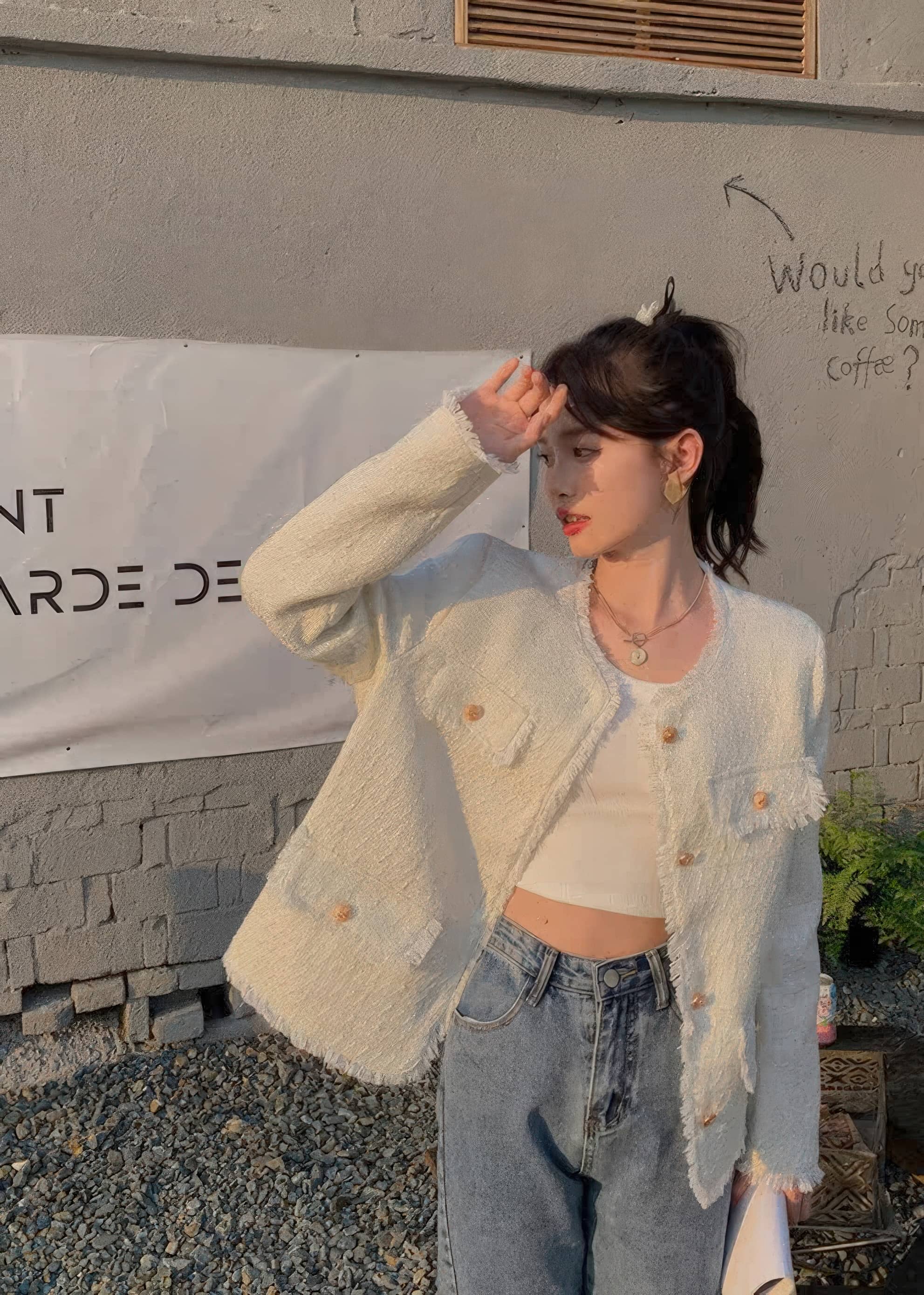 A medium close-up of a young woman with a messy updo posing on a city street. She wears an open, light blue-gray tweed jacket with raw-edge detailing and gold buttons over a ribbed white top.