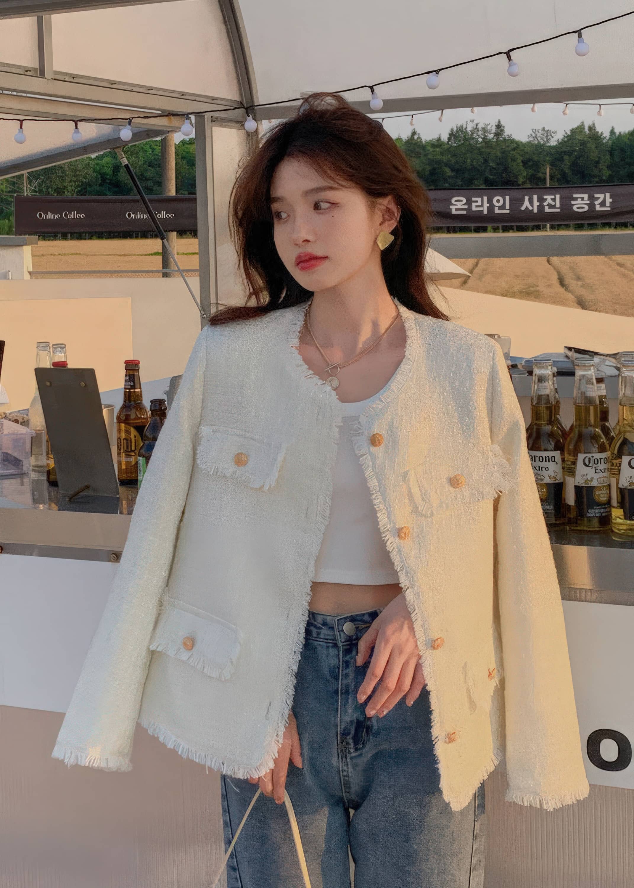 A young woman poses on a cobblestone street corner with European-style architecture in the background. She is wearing a buttoned light blue-gray tweed jacket and holding a small bouquet of blue and yellow tulips.