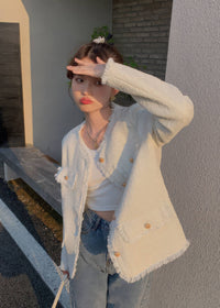 A young woman stands on an urban street holding a bouquet of tulips. She is wearing a light blue-gray tweed jacket with gold button accents and raw trim, paired with loose light-blue jeans and a yellow purse.