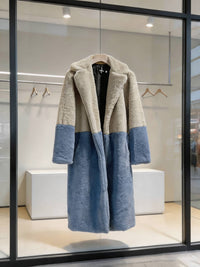 Cream grey and slate blue two-tone notch collar faux fur long coat hanging on a hanger in a store window display.