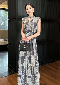 A woman poses in a full-length shot wearing a vintage-style jumpsuit with a grey and white damask print. She is standing against a dark ribbed wall, holding a small black structured handbag. The jumpsuit features a v-neckline, a matching neckband, and a wide black belt with a decorative silver buckle.