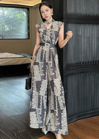 A woman poses in a full-length shot wearing a vintage-style jumpsuit with a grey and white damask print. She is standing against a dark ribbed wall, holding a small black structured handbag. The jumpsuit features a v-neckline, a matching neckband, and a wide black belt with a decorative silver buckle.