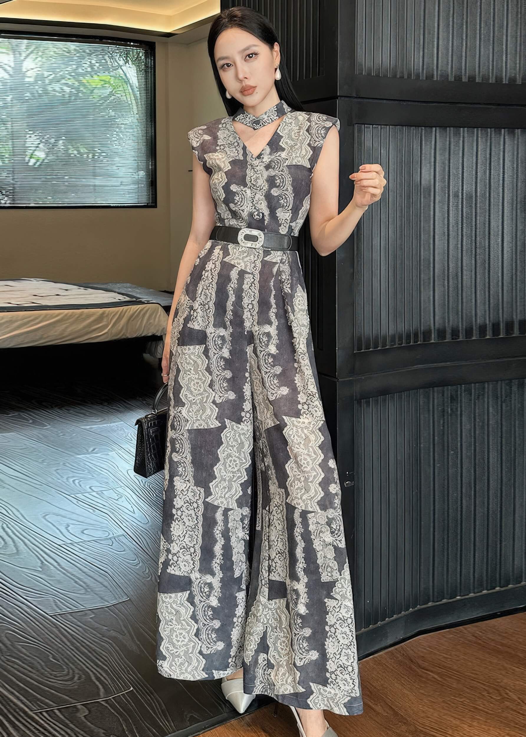 A woman poses in a full-length shot wearing a vintage-style jumpsuit with a grey and white damask print. She is standing against a dark ribbed wall, holding a small black structured handbag. The jumpsuit features a v-neckline, a matching neckband, and a wide black belt with a decorative silver buckle.
