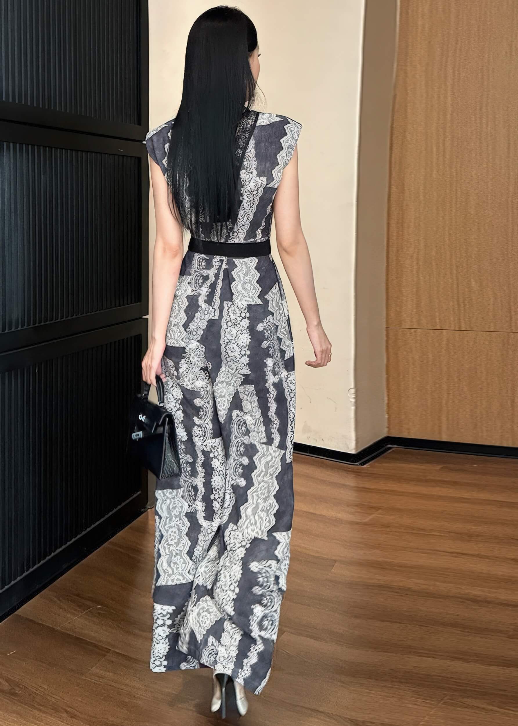 A rear view of the woman walking away, highlighting the high-waisted fit and the floor-length wide-leg silhouette of the printed jumpsuit. She is carrying a black crocodile-pattern handbag, and her long black hair falls down her back.