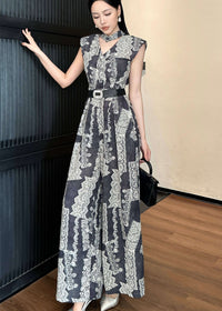 A full-length fashion shot of the woman standing with one hand resting on a dark wall and the other holding a black handbag. She is wearing pointed-toe silver heels that complement the cool tones of the wide-leg jumpsuit.
