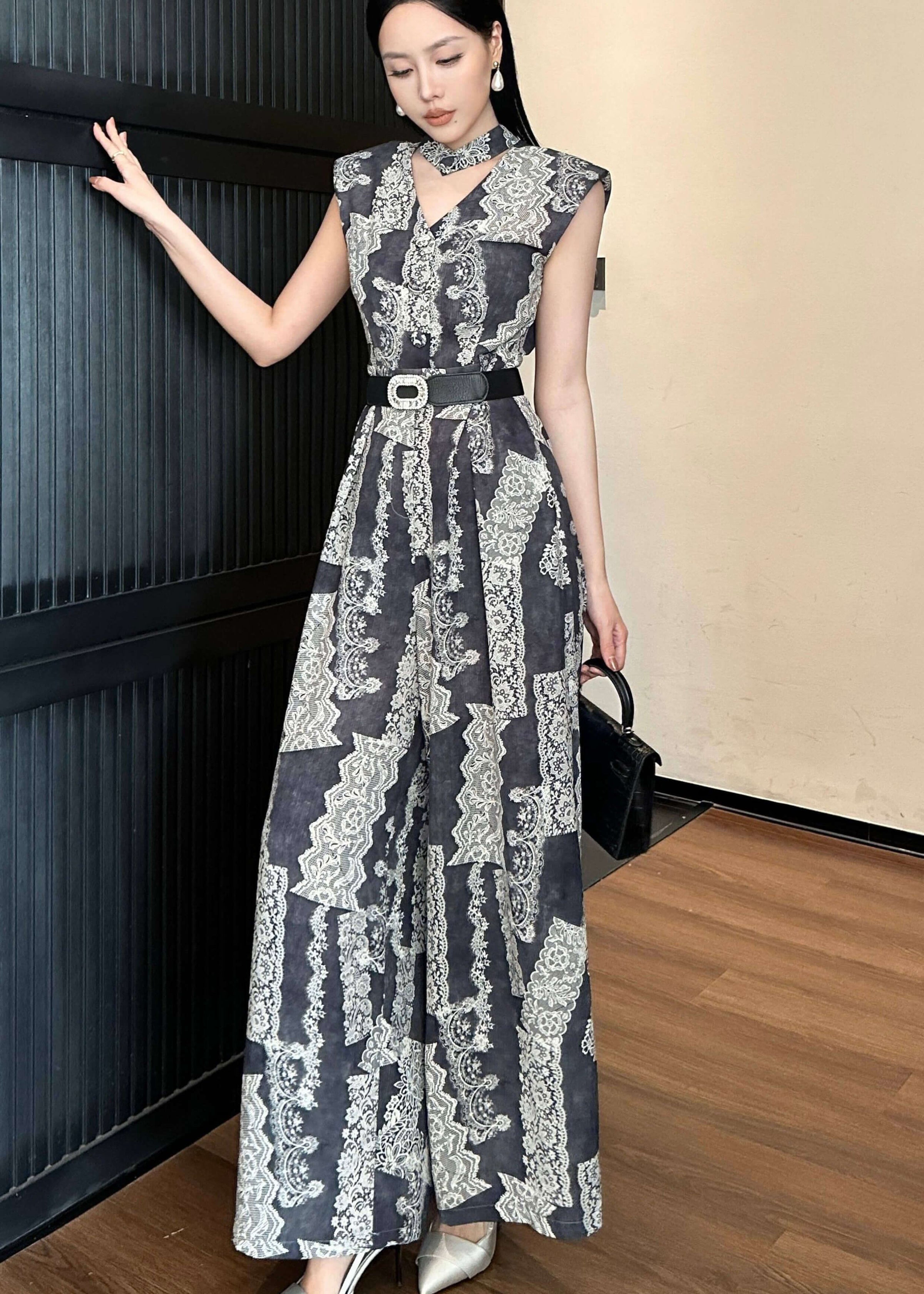 A full-length fashion shot of the woman standing with one hand resting on a dark wall and the other holding a black handbag. She is wearing pointed-toe silver heels that complement the cool tones of the wide-leg jumpsuit.
