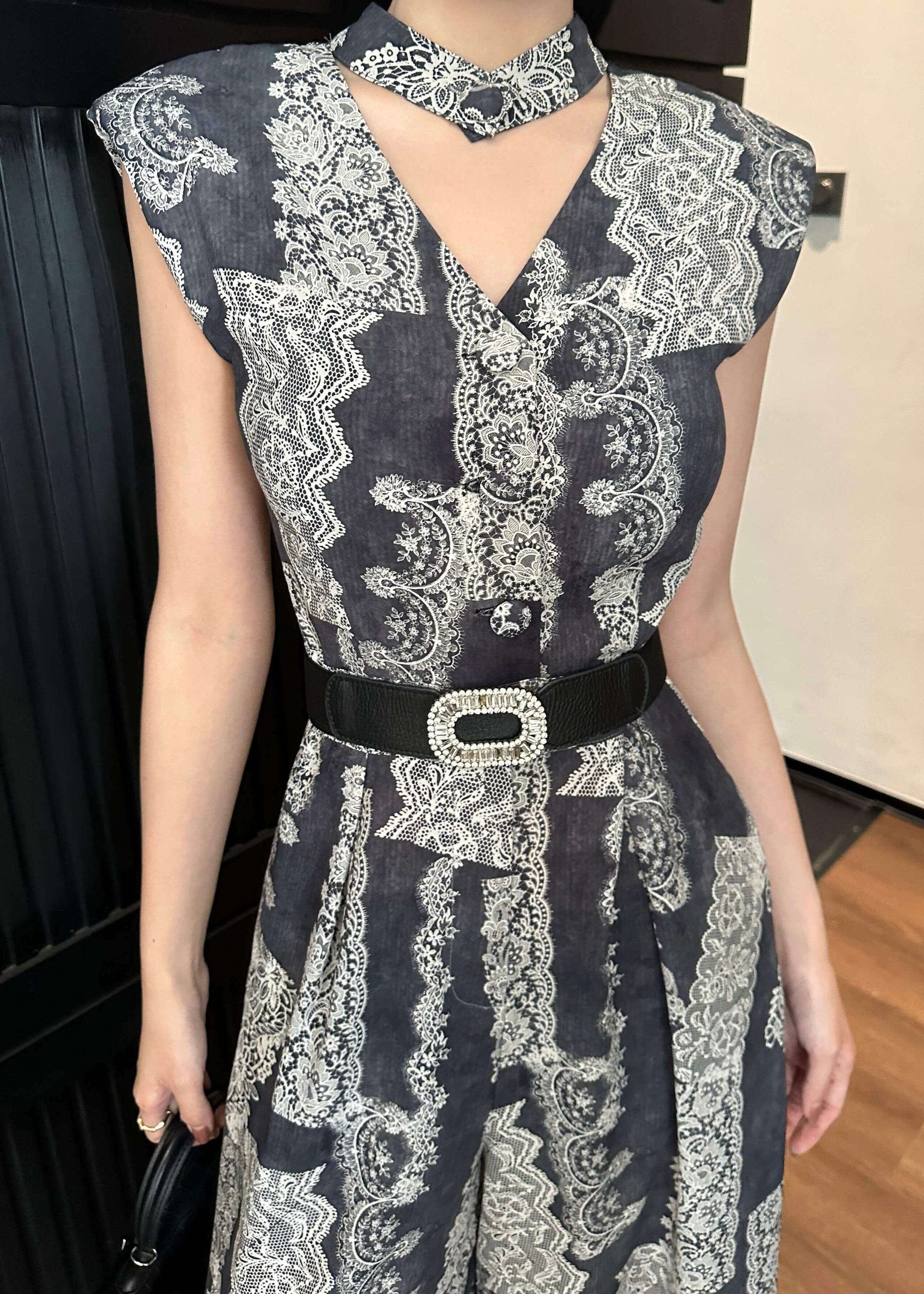 A medium close-up shot focusing on the bodice of the jumpsuit. The image details the intricate white lace-like patterns on the dark fabric, the v-neck design with a button detail, and the matching choker-style neckpiece. The decorative crystal-encrusted buckle of the black belt is prominently visible.