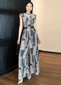 A full-length fashion shot of the woman standing with one hand resting on a dark wall and the other holding a black handbag. She is wearing pointed-toe silver heels that complement the cool tones of the wide-leg jumpsuit.