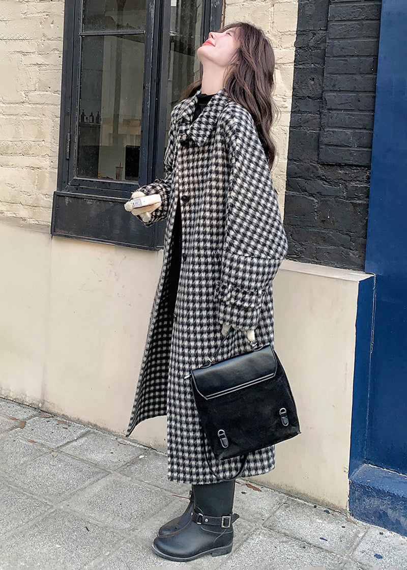A side view of the woman in a houndstooth coat and black boots standing against a brick and blue wall.