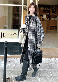 A woman smiling in an open houndstooth coat and black boots, carrying a large white scarf and a black bag.