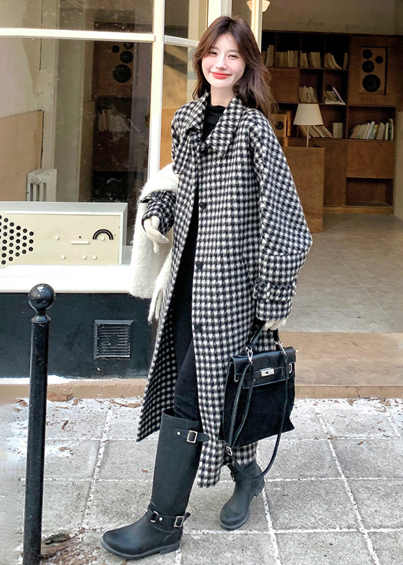 A woman smiling in an open houndstooth coat and black boots, carrying a large white scarf and a black bag.
