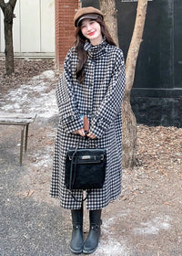 A woman in a houndstooth coat, brown newsboy cap, and black boots posing outdoors holding a black handbag.