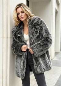Front view of a woman leaning against a wall, lightly holding closed a grey-and-black textured faux fur coat with wide lapels.