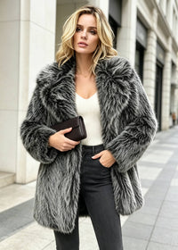 Model posing on a sidewalk in a grey faux fur coat with wide lapels, holding a black clutch purse with one hand in her pocket.