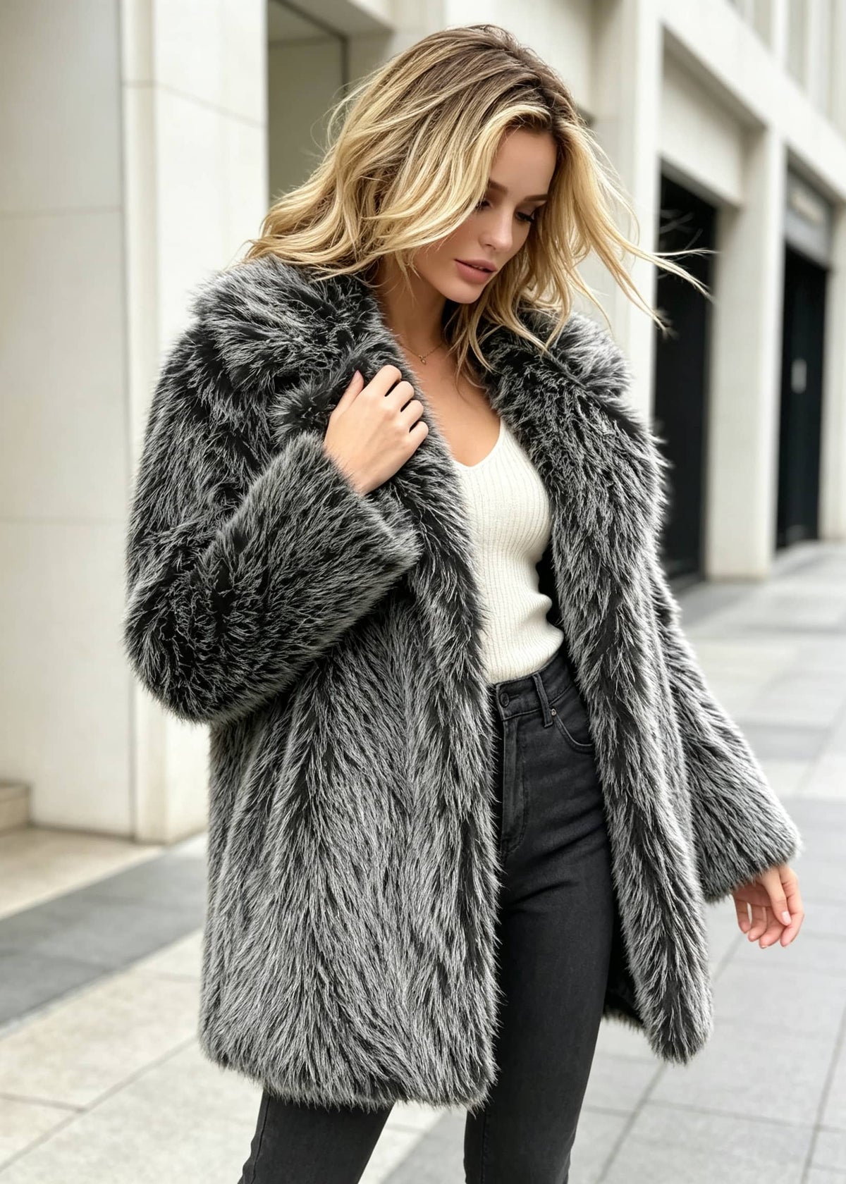 Mid-stride shot of a woman outdoors wearing a grey-and-black faux fur spread collar coat over a white top and dark jeans.