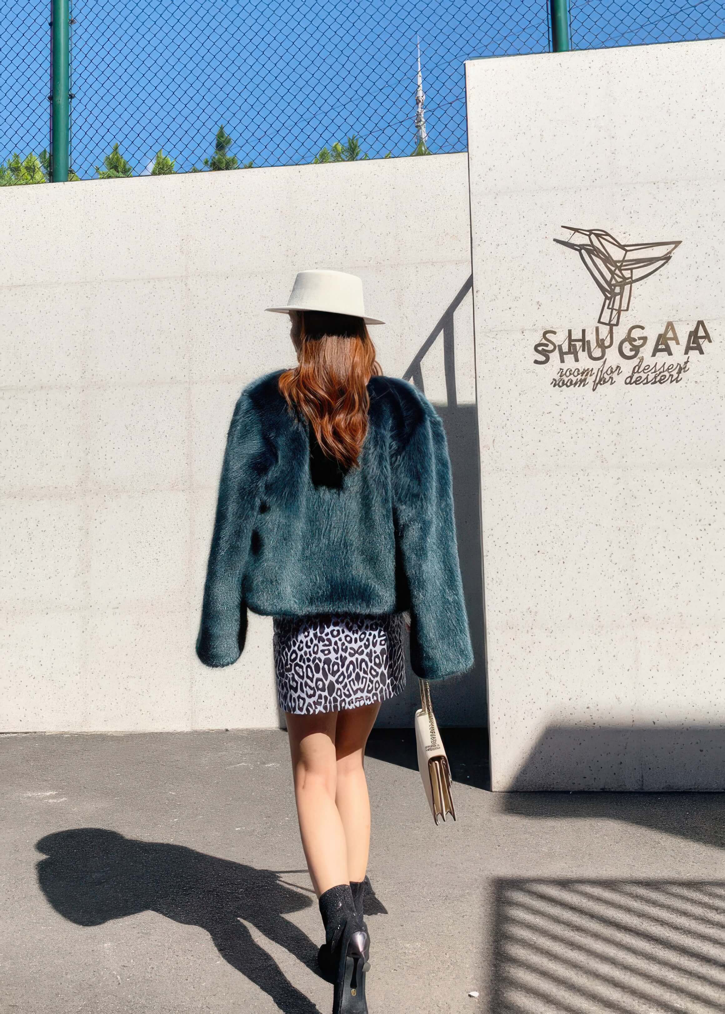  Back view of a woman wearing a dark teal green faux fur jacket and a leopard print mini skirt standing in front of a white wall.
