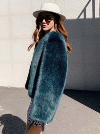 Side profile of a woman wearing a teal green faux fur jacket, a white felt fedora hat, and tortoiseshell sunglasses, showcasing the soft texture of the luxury outerwear.