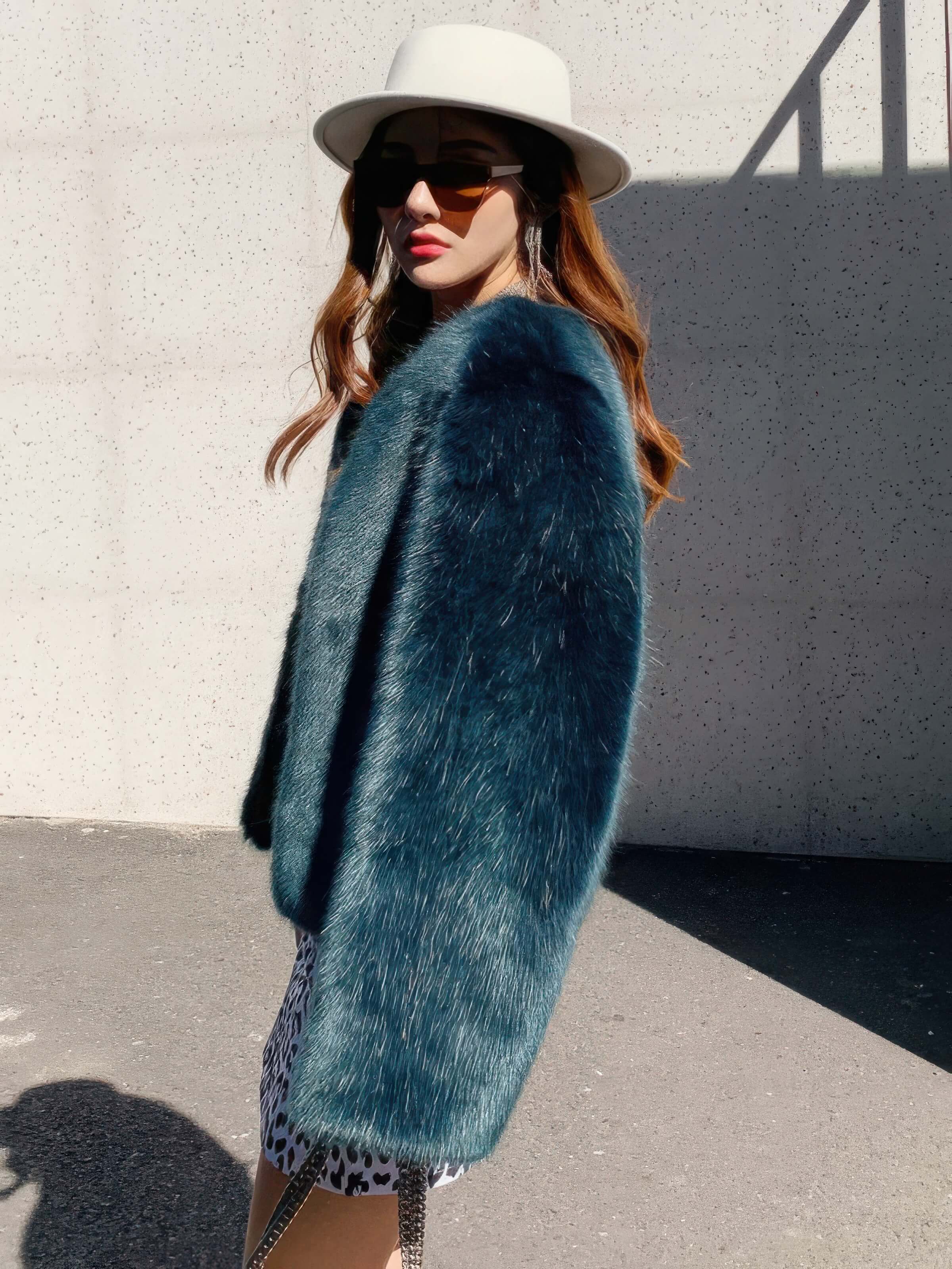 Side profile of a woman wearing a teal green faux fur jacket, a white felt fedora hat, and tortoiseshell sunglasses, showcasing the soft texture of the luxury outerwear.