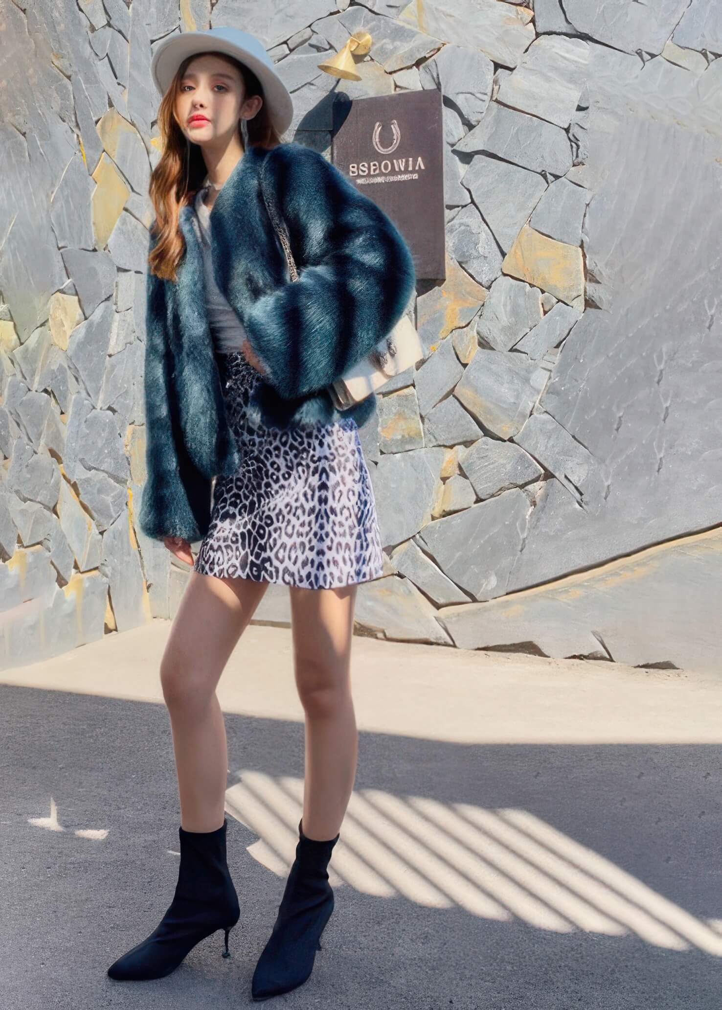 Full-length view of a woman posing in a teal blue faux fur coat, leopard print skirt, and pointed-toe black boots against a stone exterior.