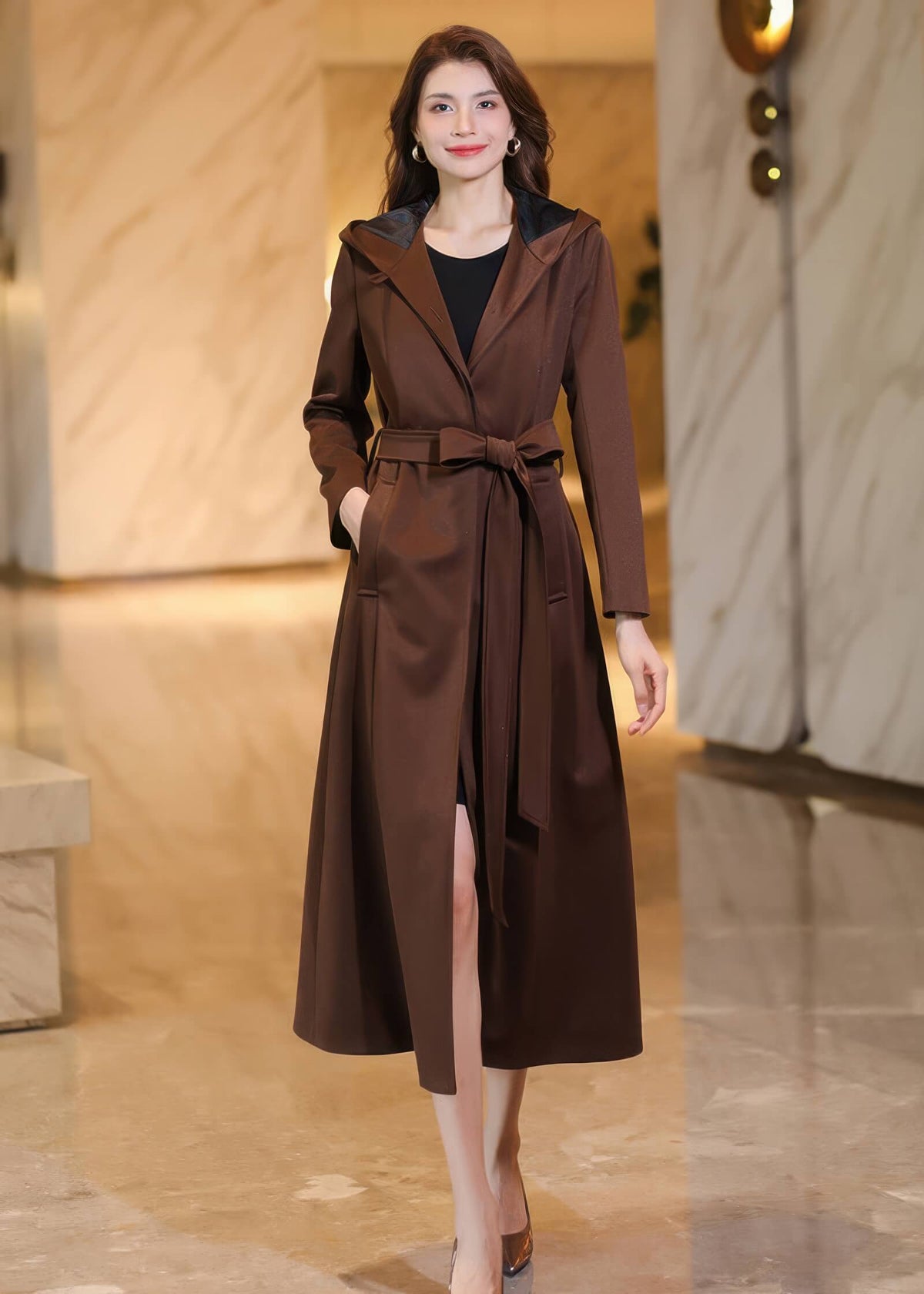 Front view of a women's long brown hooded trench coat worn open with a waist tie, styled over a black dress.