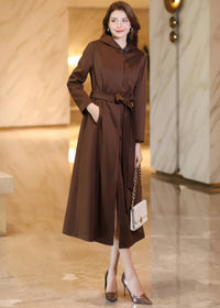 Three-quarter view of a hooded brown maxi trench coat buttoned at the neck, featuring a matching belt and side pockets.