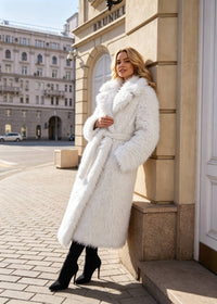 Side angle of a model leaning against a classic building column wearing a white fluffy faux fur belted coat with black heeled ankle boots.