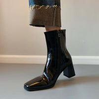 A side view of a glossy black square-toe ankle boot being worn with cuffed denim, highlighting the side zipper and sturdy block heel.