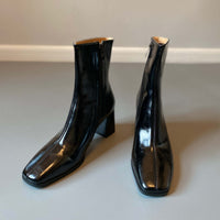 A pair of sleek, black ankle boots with a glossy patent finish, featuring a square toe and a mid-height block heel, displayed against a neutral gray background.