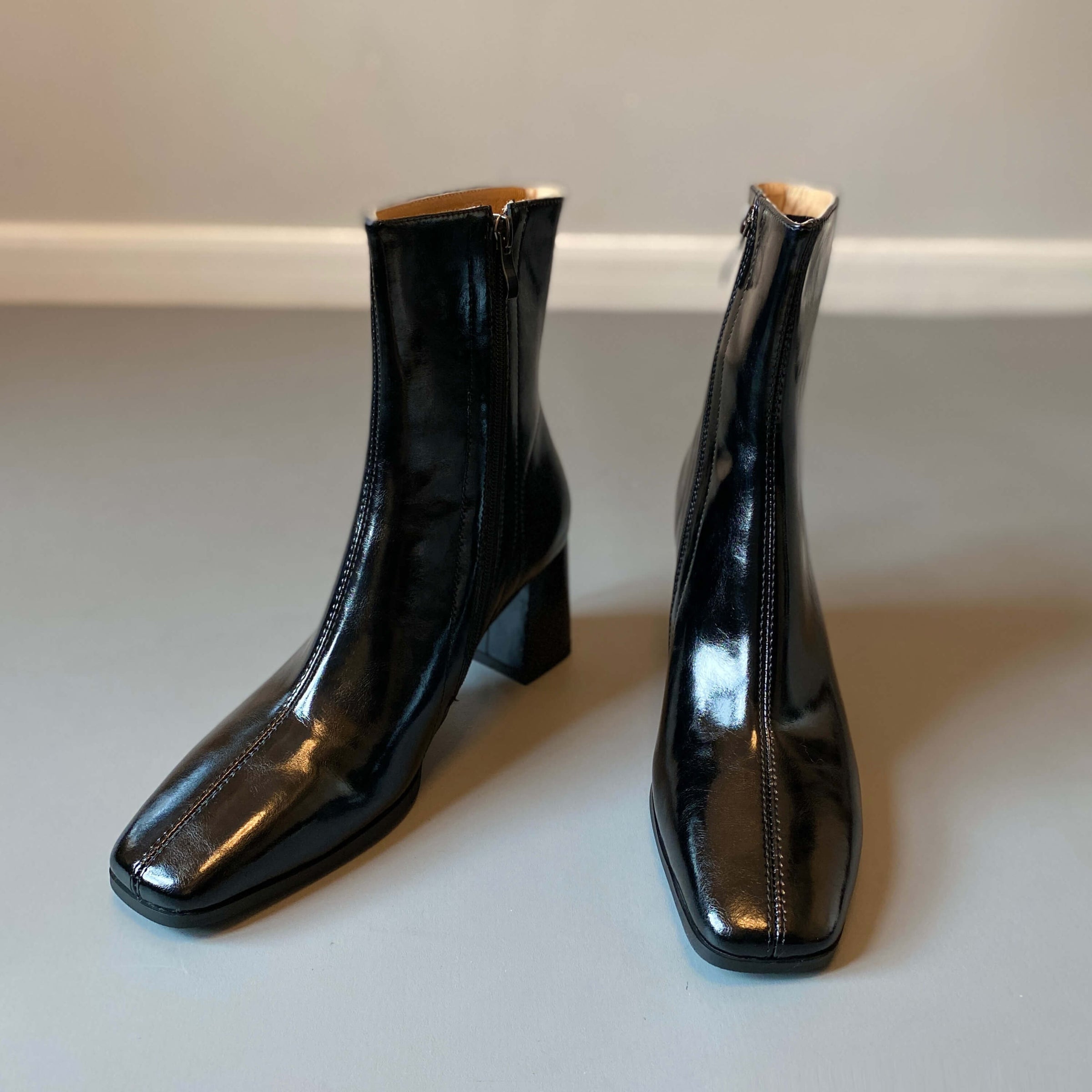 A pair of sleek, black ankle boots with a glossy patent finish, featuring a square toe and a mid-height block heel, displayed against a neutral gray background.