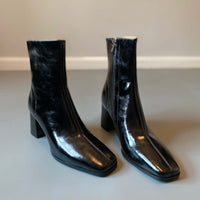 A pair of glossy black ankle boots with a square toe and block heel displayed together against a neutral gray background.