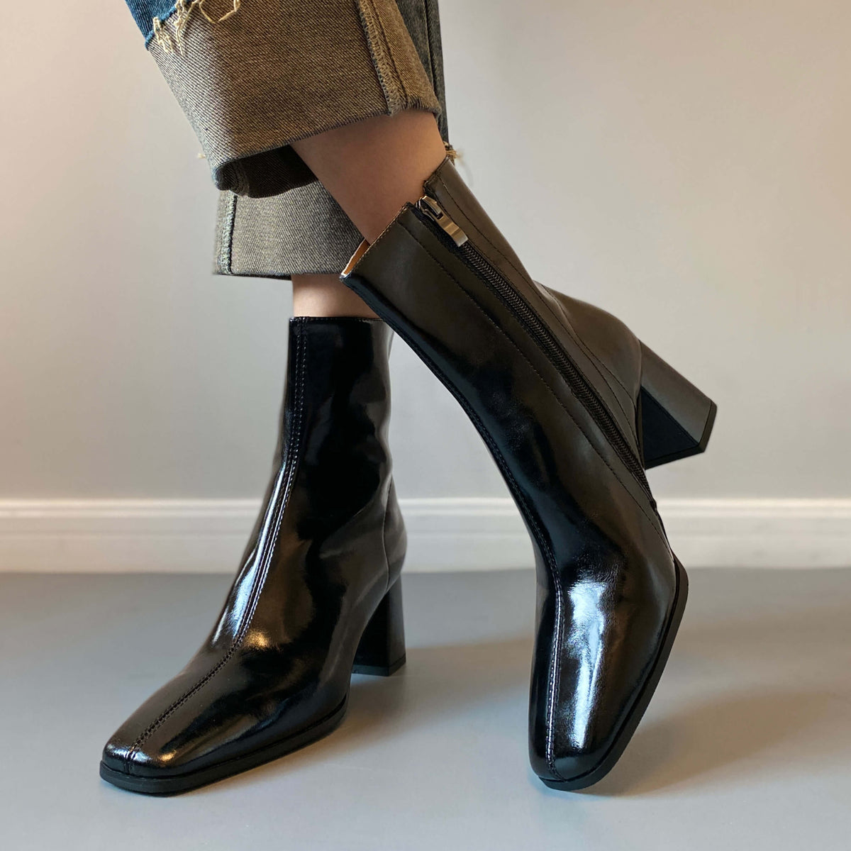 A woman posing in glossy black patent leather ankle boots, featuring a square toe and block heel, paired with cuffed blue jeans.