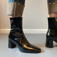 A side-profile shot of the black patent leather boots being worn with cuffed jeans, highlighting the glossy finish and the sturdy block heel.