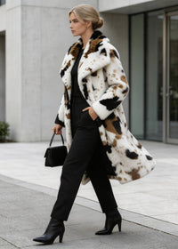  Side view of a model walking in an abstract cow spot faux fur long coat holding a black handbag on a city sidewalk.
