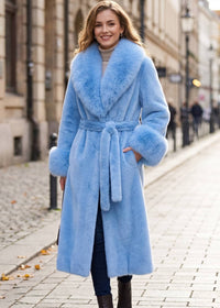A woman wearing a light blue faux fur belted coat with a large matching collar, standing on a cobblestone city street.