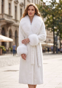A woman wearing a luxurious white fur coat with a wide collar, matching belt, and fluffy cuff details, standing in an outdoor urban setting with blurred buildings in the background.