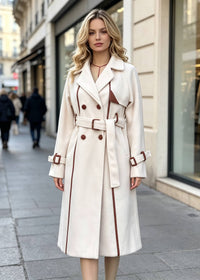 A woman stands on a city sidewalk wearing a white-colored double-breasted trench coat with brown trim and a matching belt. The background shows blurred storefronts and pedestrians.