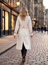 A woman viewed from behind walking down a cobblestone street wearing a long white coat with a matching belt, dark trousers, and brown ankle boots.