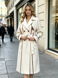 A woman stands on a city sidewalk wearing a long white-colored double-breasted coat with brown trim and a matching belt. The background shows blurred storefronts and pedestrians.