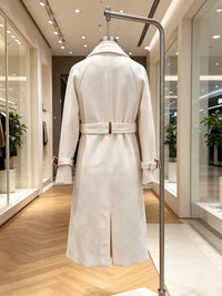 A long white-colored coat displayed on a mannequin in a boutique. The coat features a matching belt with a small brown leather accent, shown from the back view.
