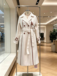 A cream-colored double-breasted trench coat with brown trim and buttons, displayed on a mannequin torso inside a modern clothing store.