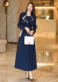 A woman posing in the Vivian Seven navy blue trench coat, tied at the waist with a matching belt. She is holding a white quilted handbag, highlighting the coat's elegant, tailored silhouette.