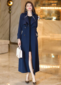 A full-length front view of a woman wearing the Vivian Seven long navy blue trench coat, worn open to reveal a black dress. The coat features cream-colored piping on the wide lapels and double-breasted buttons.