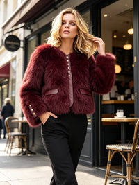 A woman stands on a city sidewalk in front of a storefront, wearing a cropped burgundy faux-fur jacket with gold-tone buttons and black pants.