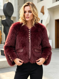 A woman wearing a cropped, plush burgundy faux fur jacket with gold-tone buttons down the front and flap pockets, paired with black pants, standing in an outdoor urban setting.