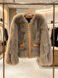 A light brown faux fur jacket with tan leather trim and gold buttons, hanging on a wooden hanger on a clothing rack inside a boutique.